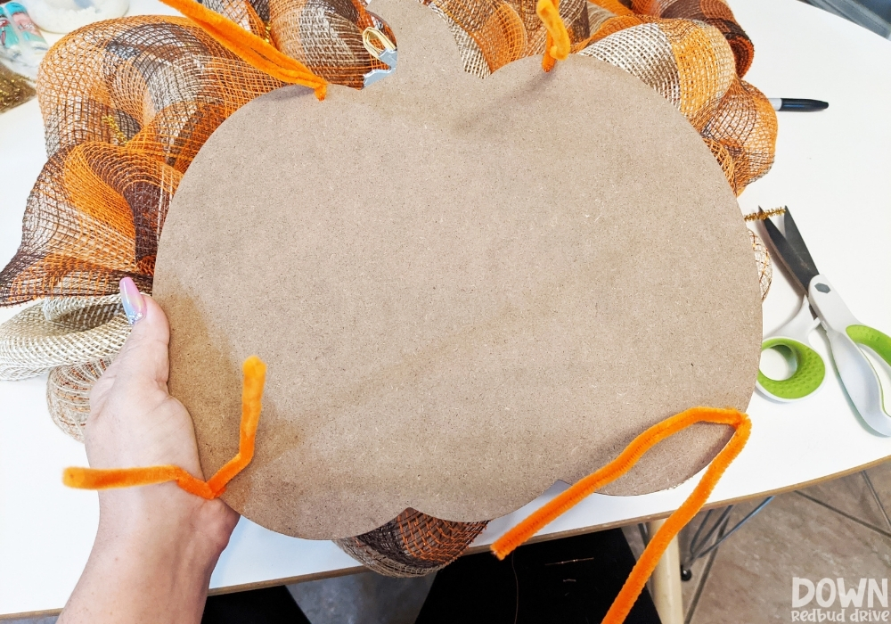 A fall pumpkin sign being attached to a wreath form with orange pipe cleaners.