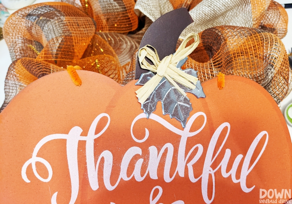 A close up of the pumpkin sign attached to a wreath form with orange pipe cleaners for the DIY Thanksgiving Mesh Wreath.