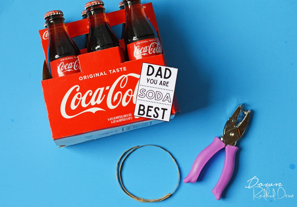 6 pack of glass coke bottles, a purple hole punch, and dad you are soda best tag, and a piece of brown raffia on a blue background