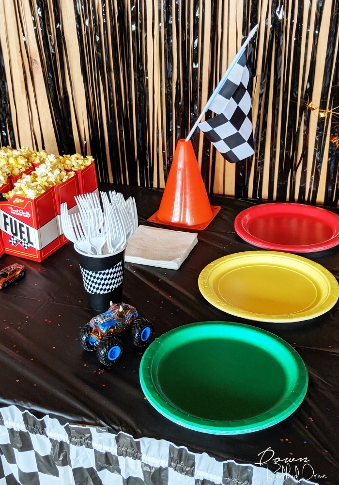 hot wheels birthday party table set up with red, green, and yellow plated, a hot wheels car, a small orange traffic cone with a checkered flag in the top of it, and white plastic forks in a black paper cup