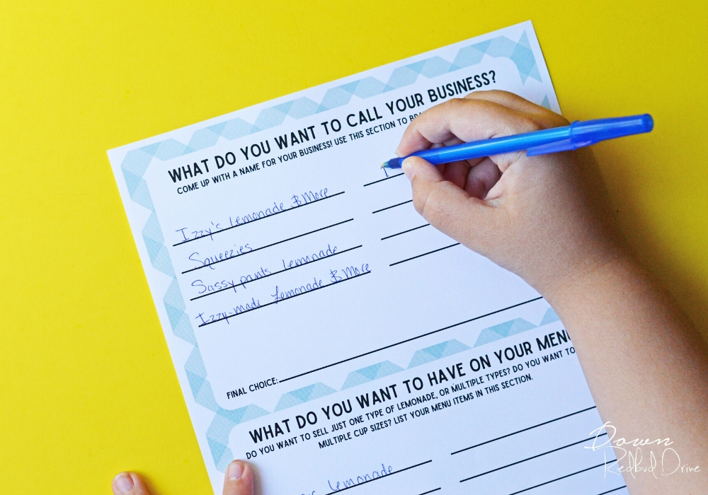 kid's hand filling out a Kid's Lemonade Stand Business Plan with a blue pen on a yellow background