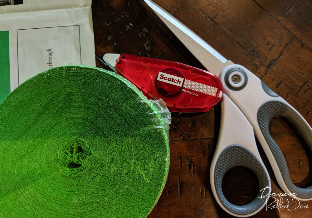 green paper streamer roll, with white scissors and double sided adhesive roller on a wooden surface