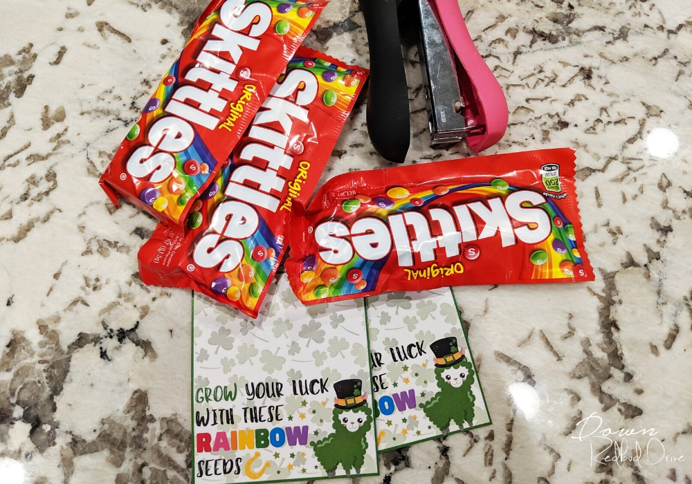 three packages of Skittles with a pink and black stapler and rainbow seeds skittles labels on a marble countertop