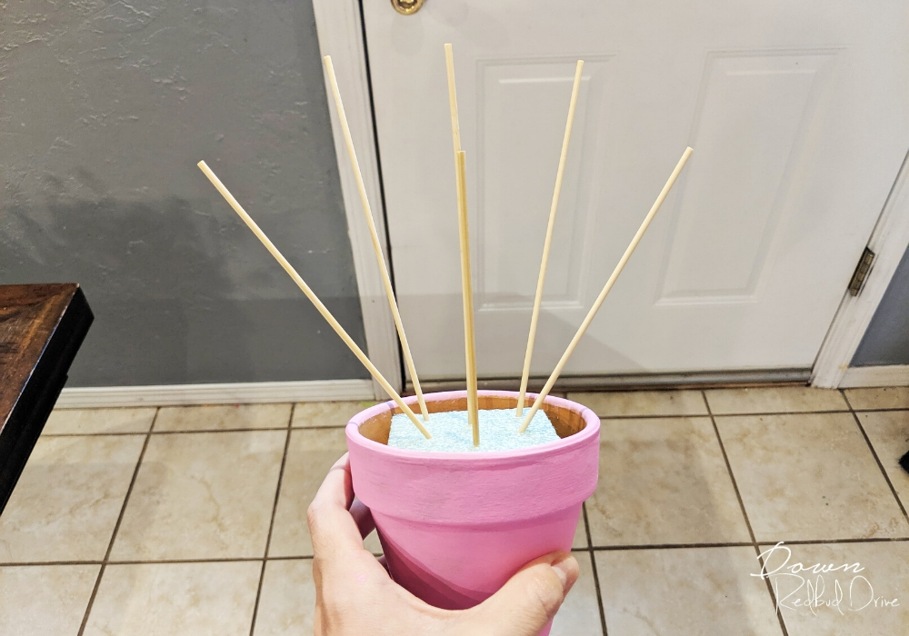 a hand holding a pink flower pot with bamboo skewers in it