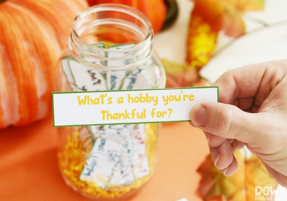 A closeup of a hand holding the question "What's a hobby you're thankful for" from the thanksgiving conversation starters game.