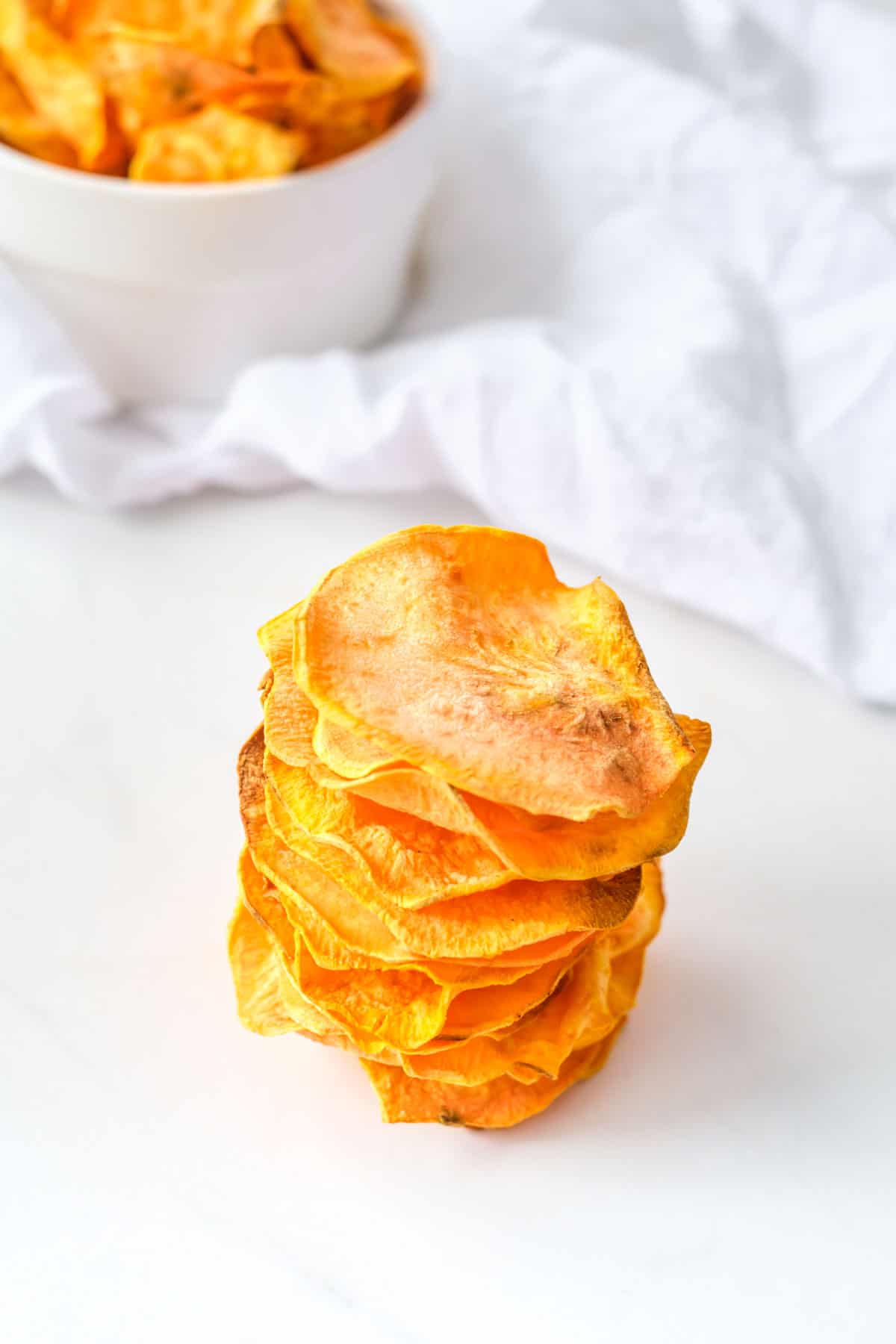 a pile of air fryer sweet potato chips on white parchment, a bowl of chips in the background.