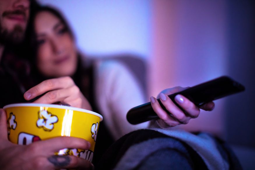 a young couple on their couch at home with a bucket of popcorn watching a movie