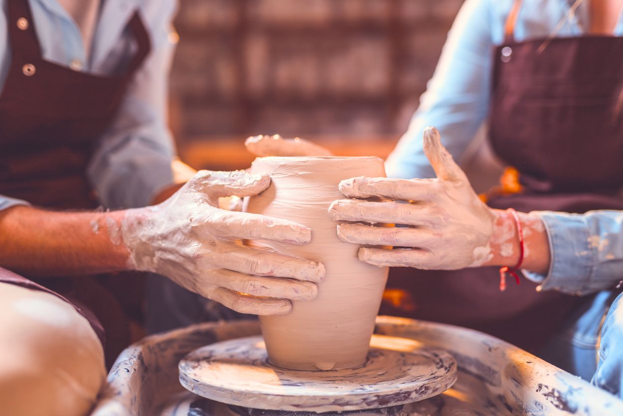 A young couple making pottery together, image is zoomed in on their hands both touching a clay pot on a pottery wheel