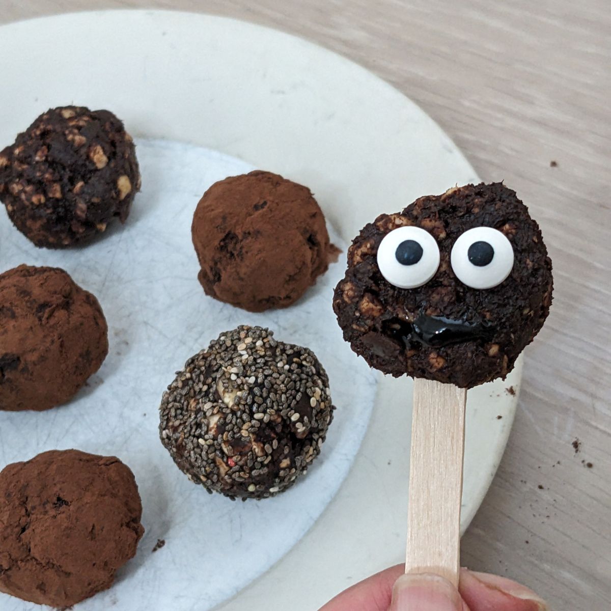 a plate of finished cocoa and orange balls, a single ball held in the foreground on a popsicle stick. Added eyes make it look like a cute monster.