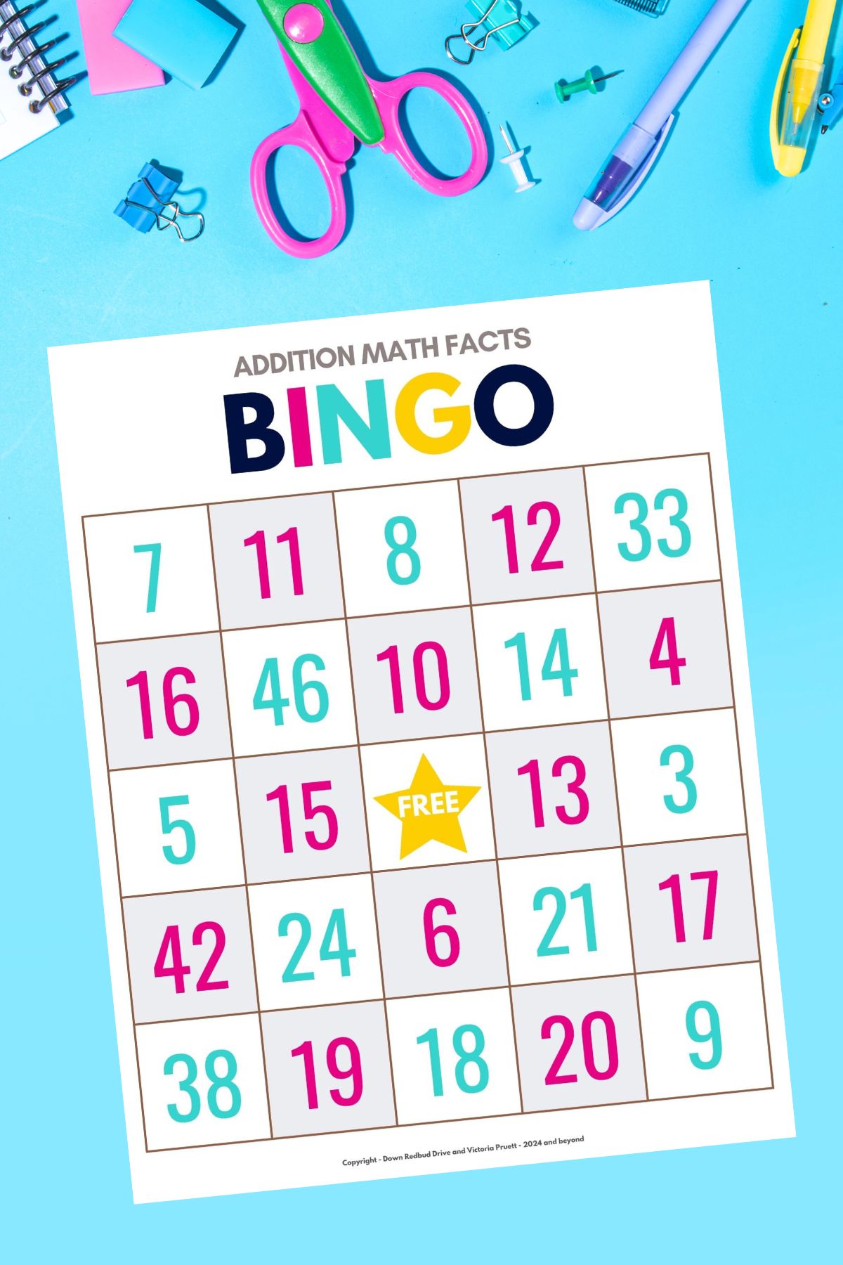 Printed bingo sheet from the addition math facts bingo game for kids free download on a bright blue background with scissors, pins, and other school supplies surrounding the page