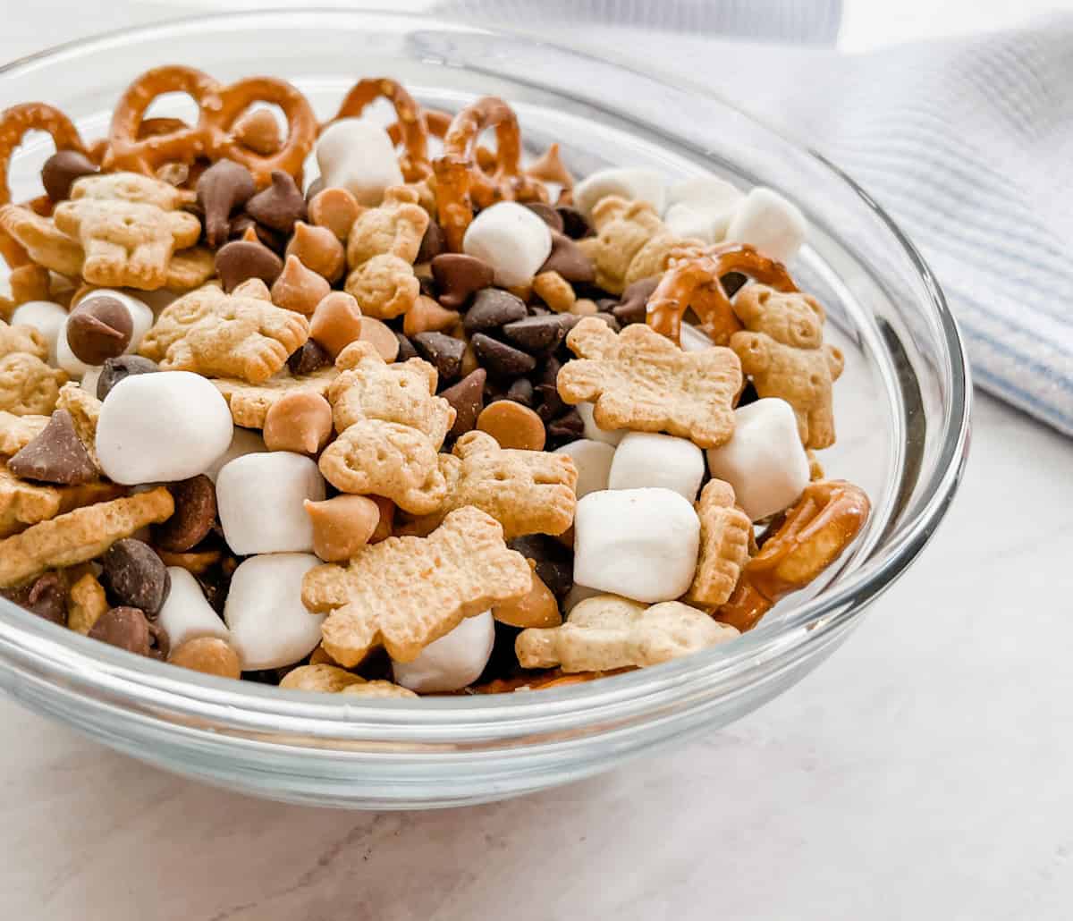 a clear glass bowl filled with s'mores trail mix made of chocolate chips, teddy grahams, pretzels, and mini marshmallows