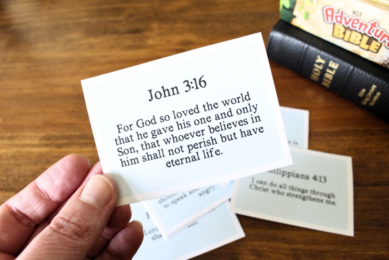 Small card cut out with a bible verse on it, john 3:16 for God so loved the world that he gave his one and only Son, that whoever believes in him shall not perish but have eternal life. The card is being held in a hand, you can see other cards in the background and 2 bibles stacks on top of each other.