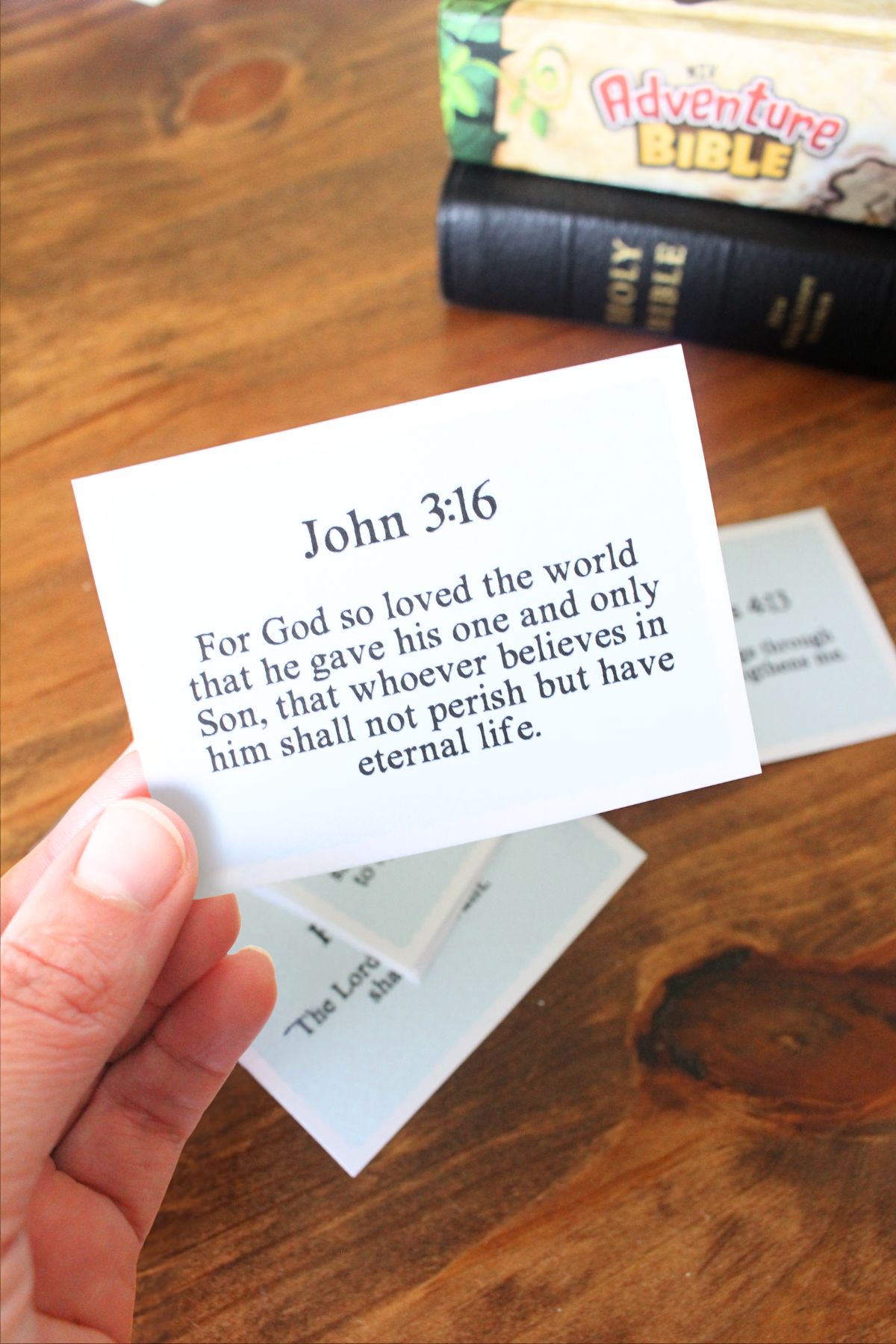 Small card cut out with a bible verse on it, john 3:16 for God so loved the world that he gave his one and only Son, that whoever believes in him shall not perish but have eternal life. The card is being held in a hand, you can see other cards in the background and 2 bibles stacks on top of each other.