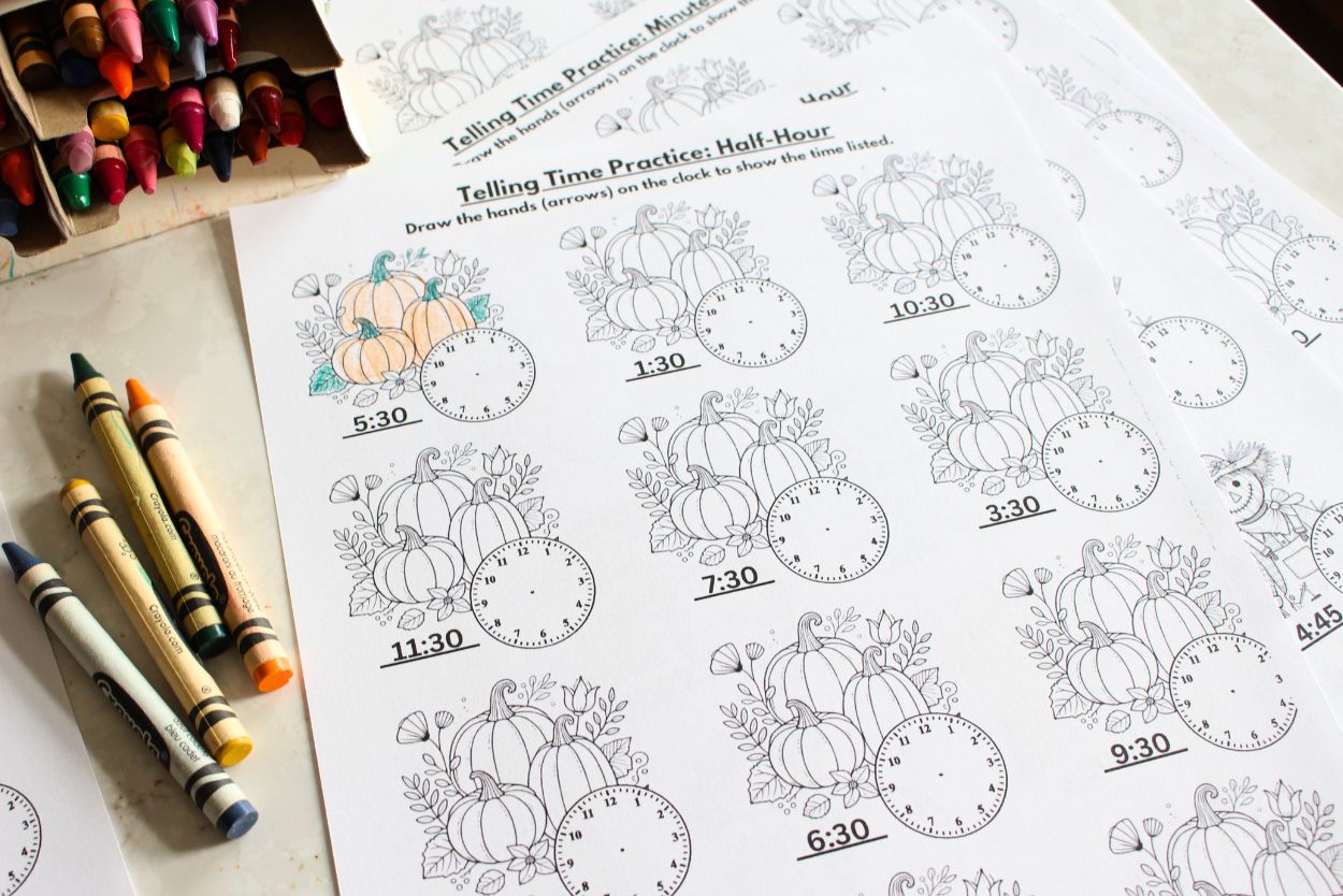 Close up of a single sheet from the fall time telling worksheet set. With pumpkins and clock faces on the sheet, the top of the paper says telling time practice, half-hours. Crayons in various colors are scattered around the paper