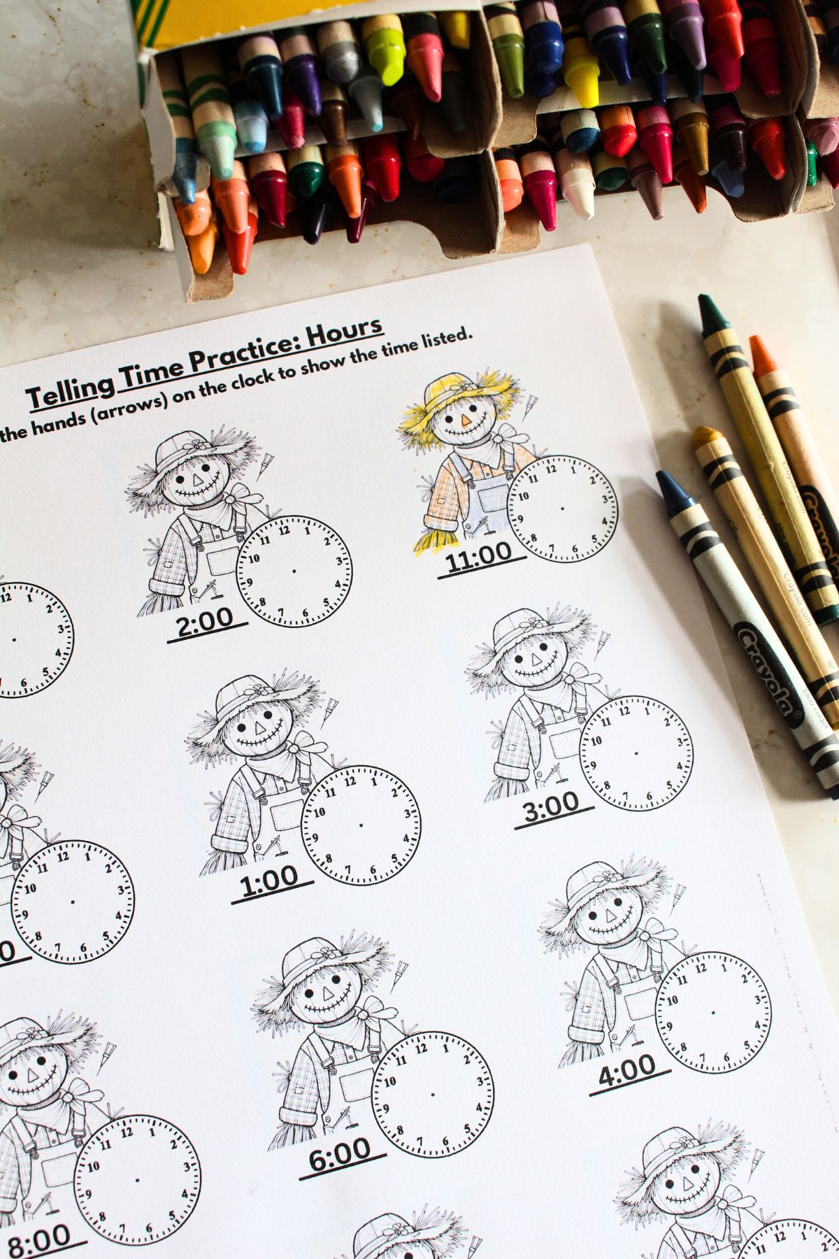 Close up of a single sheet from the fall time telling worksheet set. With scarecrows and clock faces on the sheet, the top of the paper says telling time practice, hours. Crayons in various colors are scattered around the paper