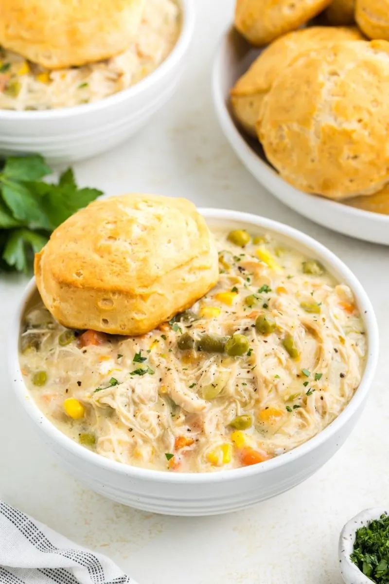 white bowl of crockpot chicken pot pie with a plate of biscuits in the background