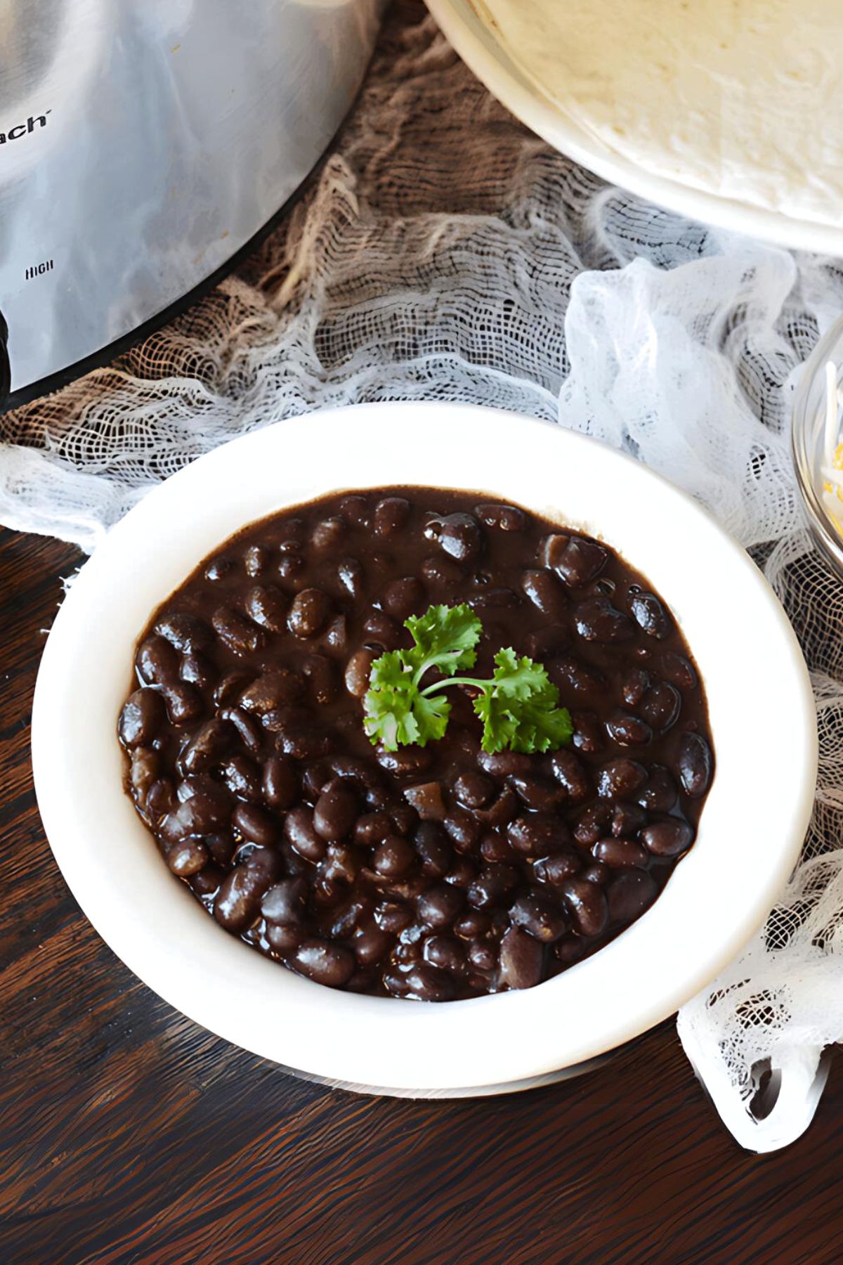 Homemade black beans in a white bowl next to a crockpot, bowl of cheese and pile of tortillas